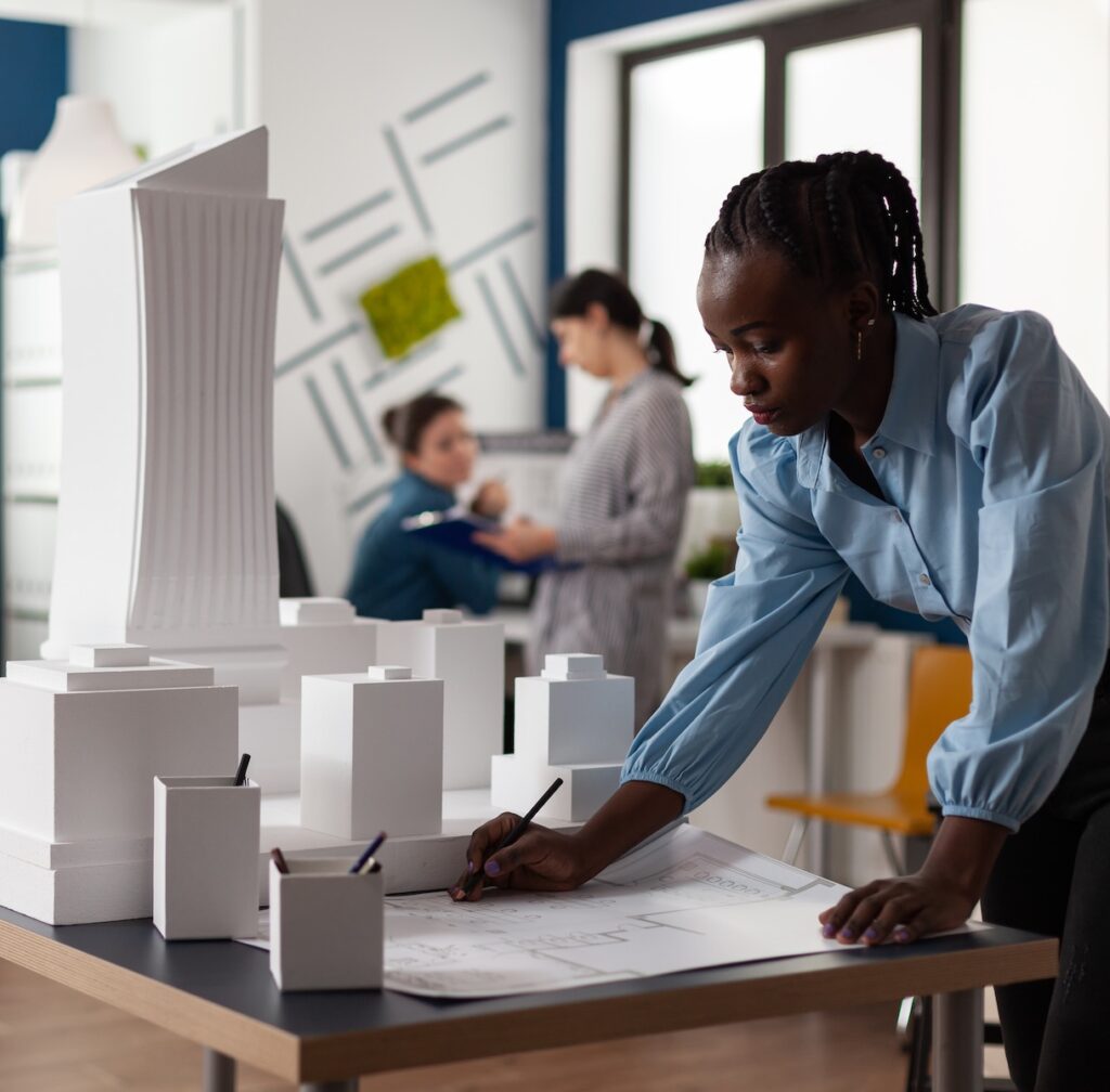 African american business woman at architectural office working on blueprint at desk. Young constructor engineer designing building model maquette in professional environment
