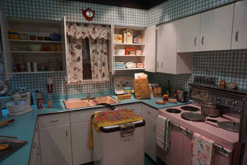 A recreated 1950s kitchen with built-in white cabinets, checkered tile walls, a pink stove, and floral curtains