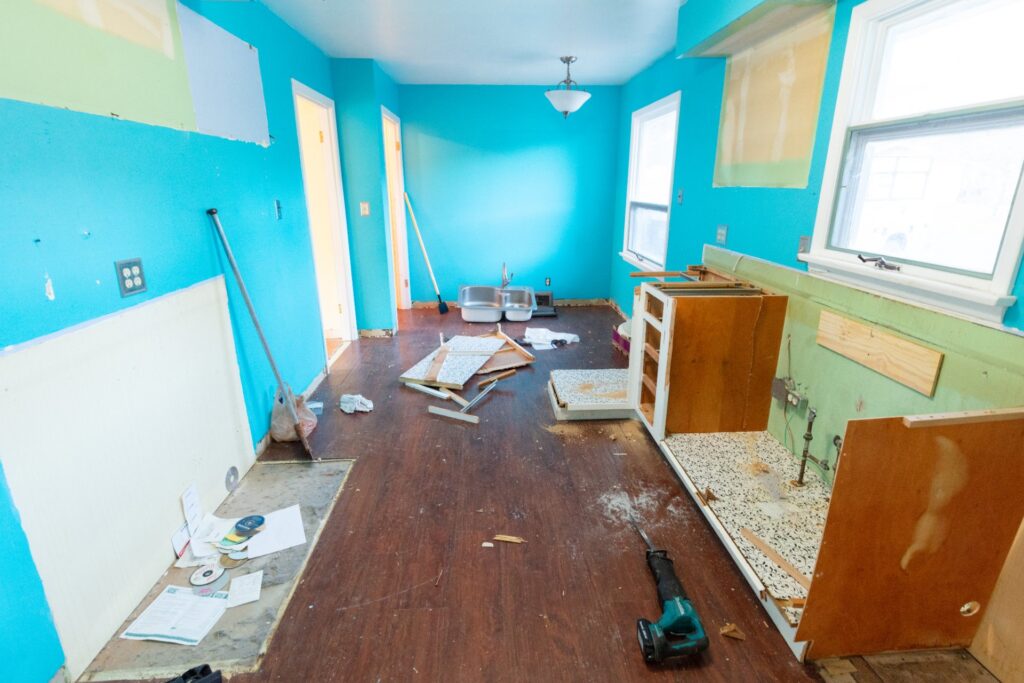 A kitchen stripped down mid-demolition with cabinets torn out, a bare wood floor, and debris scattered across the room