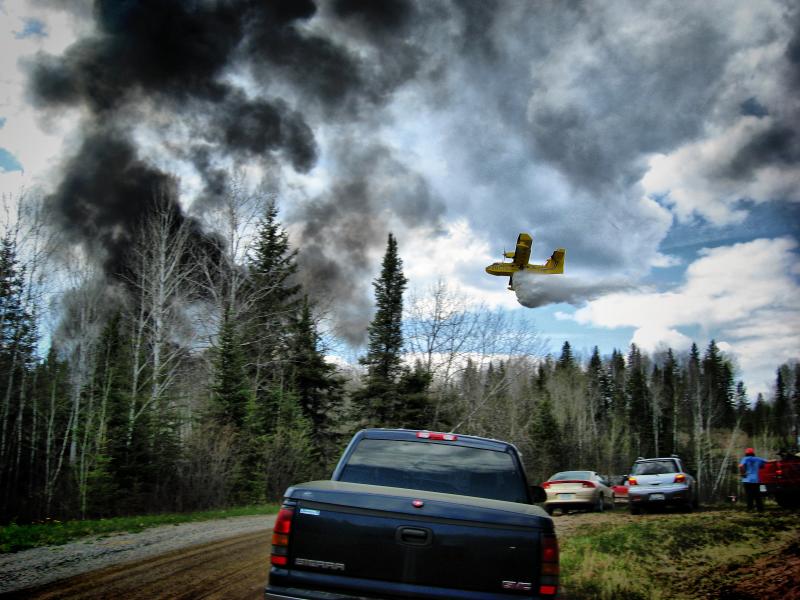 A yellow water bomber aircraft flies low over spruce trees beneath dark smoke near Thunder Bay, Ontario